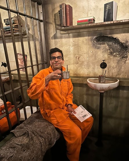 Dressed in an orange jumpsuit, Amma lifts a tin cup, sitting on a narrow bench in a cell, on a filthy blanket, everything is dirty