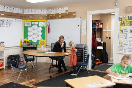 The interior of a bright, tidy classroom, with peach-painted walls, white board and bright white, green and yellow posters, and what looks like a circle of desks. An adult woman leans toward a young girl.