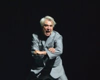 Byrne in a grey suit performs with an expressive face and dynamic pose