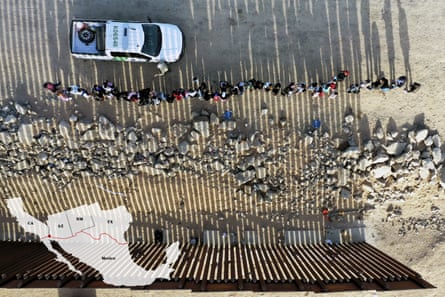 aerial view of line of people next to white car and a metal fence, with a map of mexico and the US border states superimposed on it