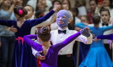 A man in evening shirt, bow tie and face mask dances with a women in purple leotard at a ballroom competition