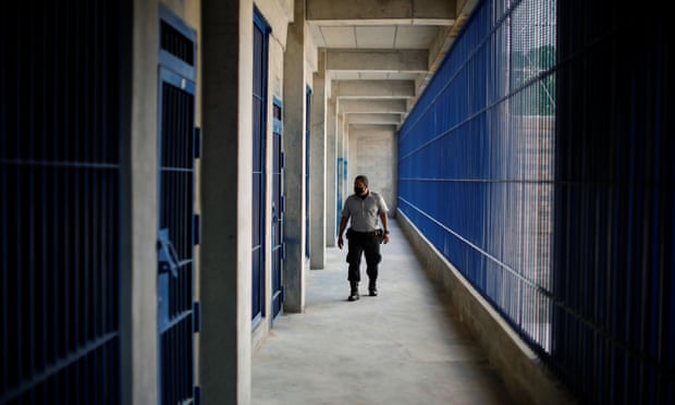 A prison guard walks down a hallway of prison cells.