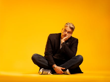 Iain Lee sitting cross-legged on the floor wearing a black suit and converse type trainers in a room bathed in bright orange light