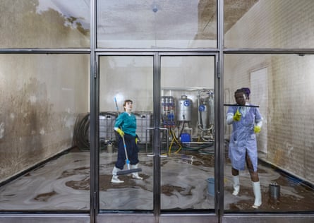 A woman uses a squeegee to clean a window as another woman also inside the room stands holding a flat mop. Boiler-type metal equipment is behind them.