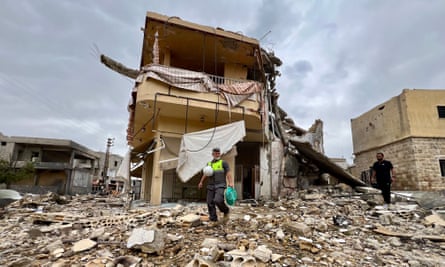 A house destroyed by an Israeli strike: its frontage and roof have been blown off and rubble is strewn all around. A man in a fluorescent vest picks his way over the debris