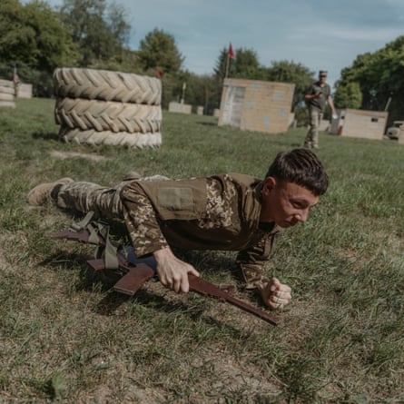 A male cadet in camouflage lies on grass holding a wooden replica rifle. He is raising himself on his elbows.