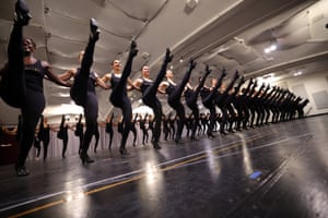 A line of dancers link arms. All have one leg in the air