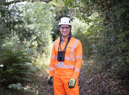 A man in an orange HS2 work uniform and white hard hat stands for a picture on a wooded track.