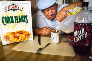 an older man in a blue shirt and blue flat cap pours milk into a mug of cereal. a box of corn flakes and a bottle of cherry soda sit beside him on the table