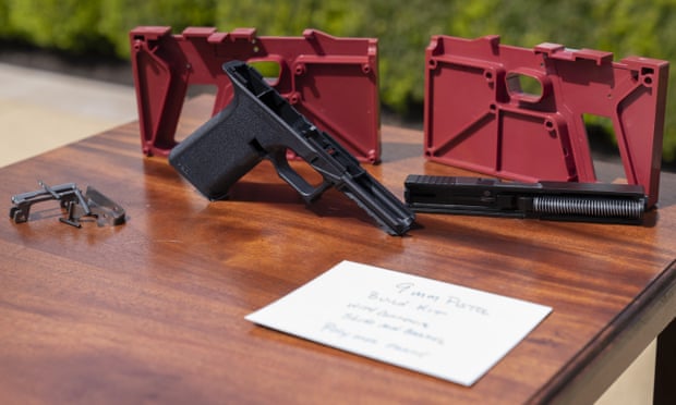 A red gun mold stands on a table next to the gun made from it. A blurred notecard in the foreground reads "9mm pistol build kit".