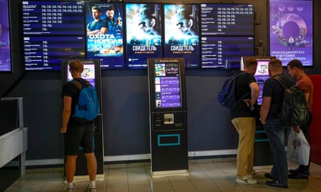 Tinerii stau la automatele de bilete într-un hol de cinema puternic iluminat, cu afișe de acțiune pe perete