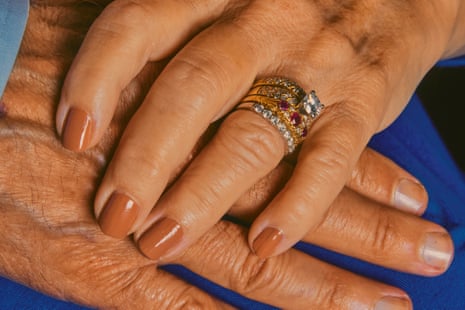 A woman’s manicured hand has four rings on the ring finger. It rests on top of a man’s hand