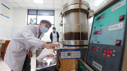 A man in a lab coat and face mask checks a container of liquid next to a screen with numbers on it in a hospital room