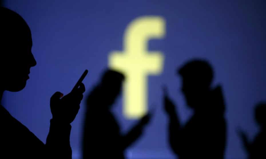 Silhouettes of mobile users are seen next to a screen projection of Facebook logo
