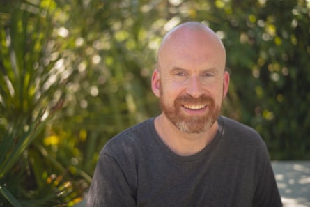Matt Haig wearing a dark-grey T-shirt, sitting in front of some bushes