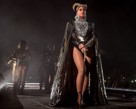 Beyoncé in an ornate black and silver cape and headdress stands center stage with backup dancers behind