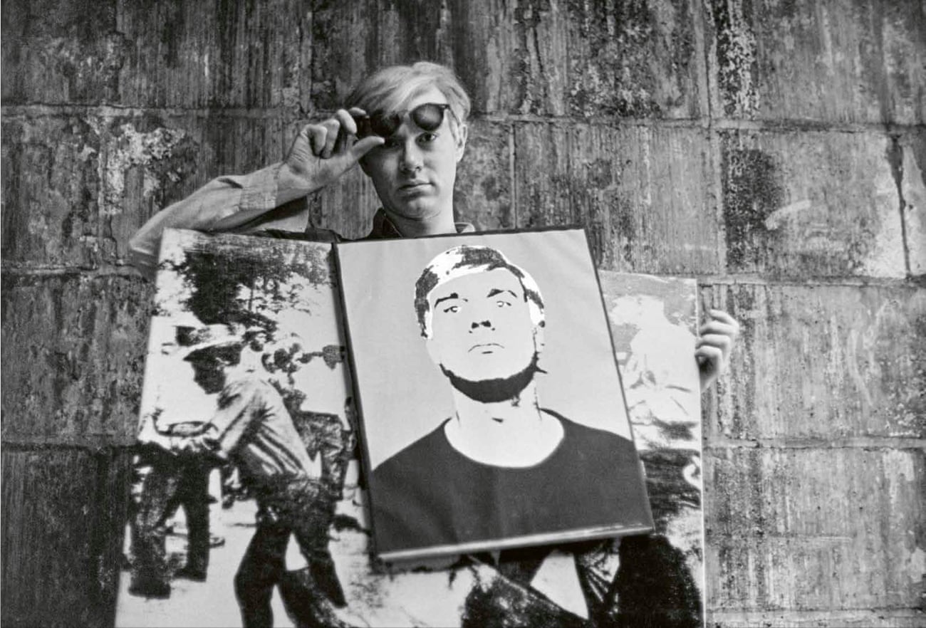 Andy Warhol wearing a sandwich board featuring some of his pictures, and holding up his sunglasses so you can see his eyes