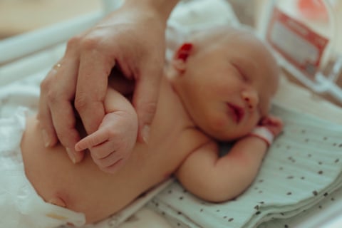 A two-day-old baby with eyes closed holding an adult’s finger.