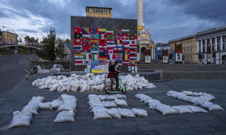 Mujeres que conducen una motocicleta a través de Maidan Square en Kyiv, pasando sacos de arena con la palabra 