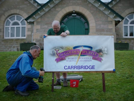 Two men are setting up a sign reading ‘The Golden Spurtle’ on a grassy hill outside the front of a stone building.