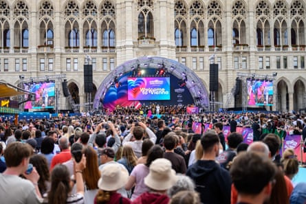 Back of crowd in foreground looking at stage with screen lit in Eurovision 2026 colours with grand building as backdrop