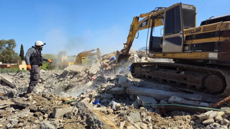 A rescue worker stands next to a digger atop a pile of rubble.