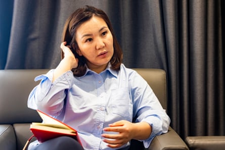 A young Mongolian woman brushes her hair behind her ear, sitting on a sofa with a red book on her knee