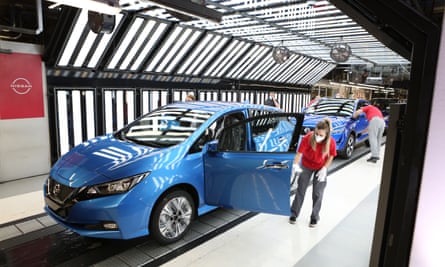 A worker in a brightly lit part of the production line attending to something on the open door of a modern looking blue hatchback