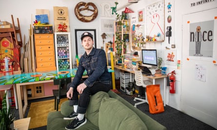Jaydon Rowbottom, with two round earrings and wearing a baseball cap, long-sleeved shirt and trainers, sits on the back of a sofa in an art studio crowded with shelves, a table, sketches pinned on the walls and a computer on a shelf