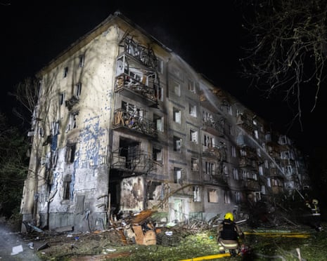 A charred and crumbled residential building in the dark, with firefigters spraying hoses at it