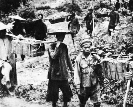 B&W photo showing porters on the Ho Chi Minh trail carrying boxes tied to canes that are carried on their shoulders.