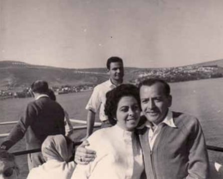 Black and white photo of a man and woman hugging with a body of water in the background and land on the other shore. He wears a buttoned-up cardigan and collared shirt. She is wearing a white long-sleeved top with a high neck