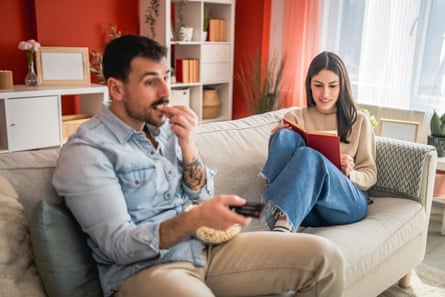 man sit on sofa watch movie and eat popcorn while woman read a bookfocused adult man sit on sofa watch movie and eat popcorn, while woman in the background read a book