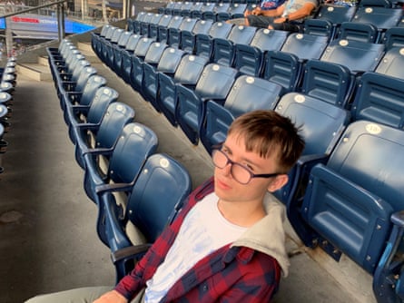 Tom Parfett looks at the camera above the rim of his glasses as he sits along a row of empty seats at a sports stadium