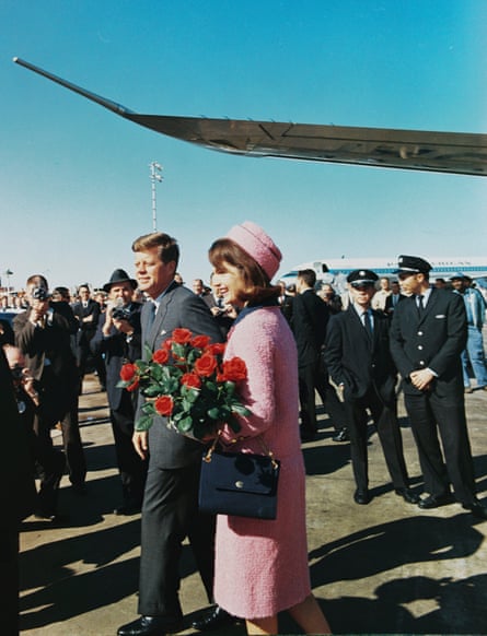 Jackie Kennedy in a fashionable pink dress and matching hat, standing next to John F Kennedy, who is wearing a suit, on an airport runway next to a plane