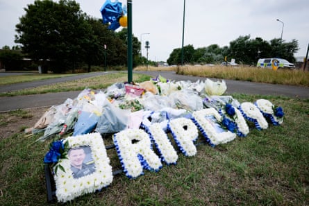 Floral tributes at the road junction. White flowers spell out RIP and 999, alongside flower-framed pictures of the two police officers. Further bunches of flowers are laid behind on the grass.