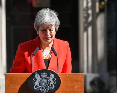 Theresa May stands outside Downing Street behind a podium and microphones, looking on the verge of tears; she is wearing a bright red jacket and chunky necklace.