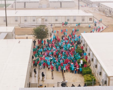 Aerial view of dozens of people in all-red or all-teal jumpsuits standing in a courtyard between two two-story industrial buildings.