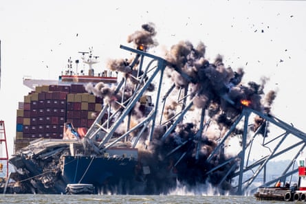 Black smoke erupts from points on a section of bridge hanging precariously off a huge container ship, which can be seen behind it
