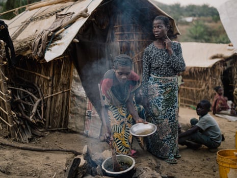 Two women outside a straw hut. One is serving food into a bowl from a pot on a fire.