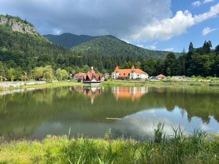A scenic view of a mountain lake surrounded by peaks and greenery with a couple of traditional buildings on the shore.