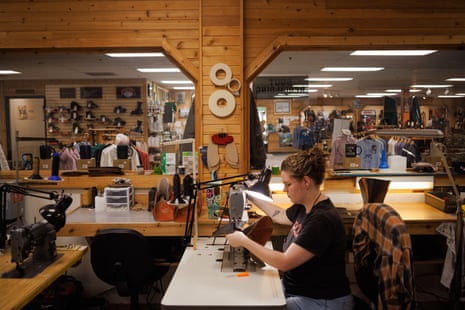 Woman sits at table holding a boot in a brightly lit room with wood paneling, with a store on the other side of glass.