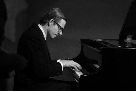 A man with slicked-back hair and glasses, wearing a dark suit