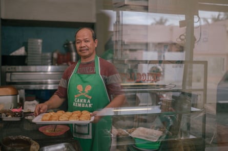 An Indonesian man in a green apron holding a tray of doughnuts in a kitchen