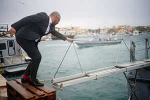 James Cleverly standing on a wooden step on a pier and holding onto a gangway that is being extended out to him from a boat