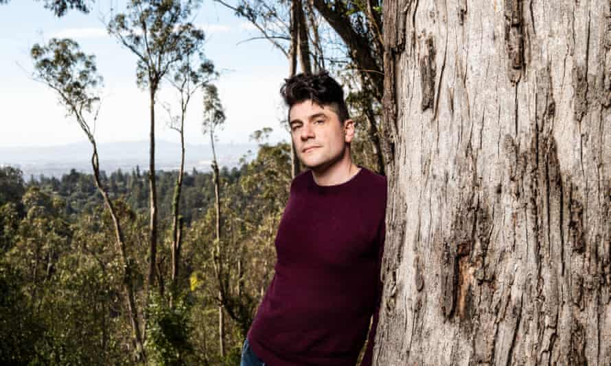 David Jay in a mauve top leaning against a tree