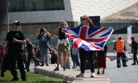 A young woman in sunglasses standing on a short wall next to a lawn holds up a Union Jack flag while being photographed on a woman’s mobile phone