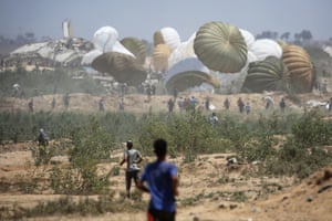 People run across dry, sandy land rush towards an area where white, green and brown parachutes are landing