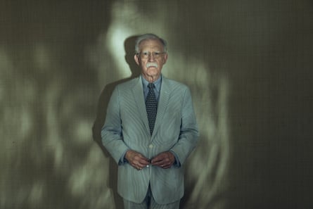 John Bolton standing against a wall wearing a pale suit, looking at the camera