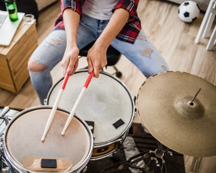 A shot of a young woman playing drums in her apartment. She is dressed in a plaid shirt, sits at the drums